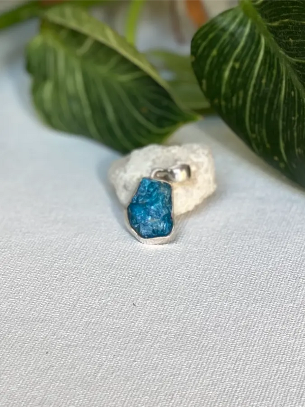Silver pendant with a blue rough Apatite stone on a light gray background with green leaves.