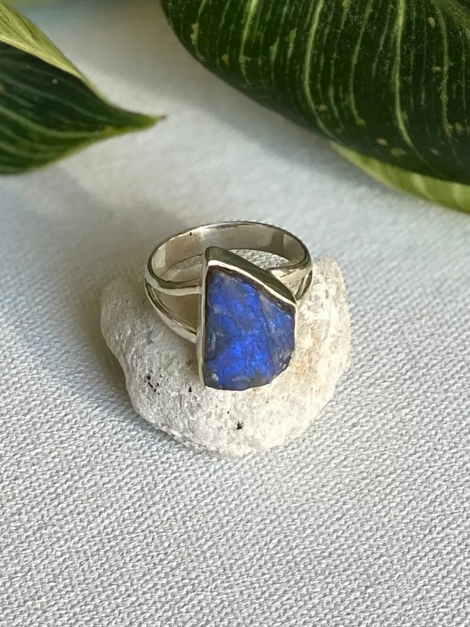 Silver ring with a blue rough labradorite stone on a white surface with green leaves in the background
