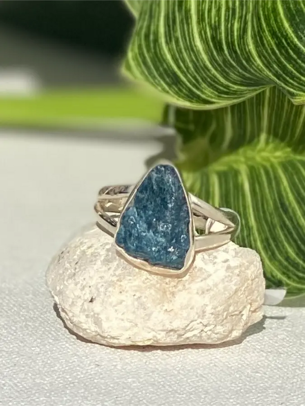 Silver ring with a blue apatite stone on a white surface with green leaves in the background