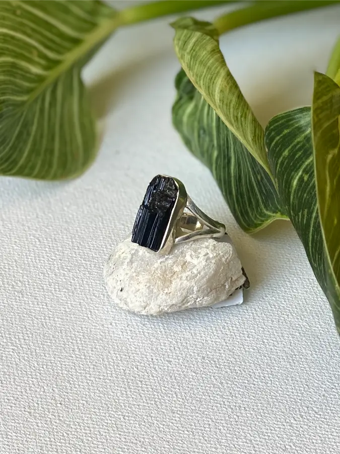 Silver ring with a black rough tourmaline stone on a white surface with green leaves in the background