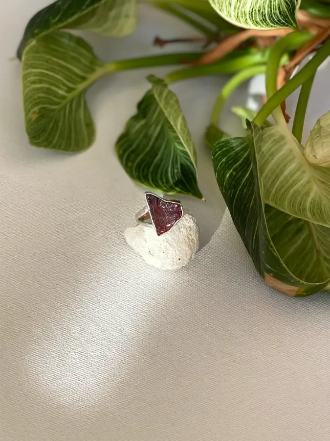 Ring with a red rough garnet stone on a beige surface with green leaves in the background