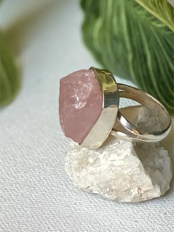 Ring with pink raw Rose Quartz gemstone and Sterling silver band on a white surface with green leaves in the background