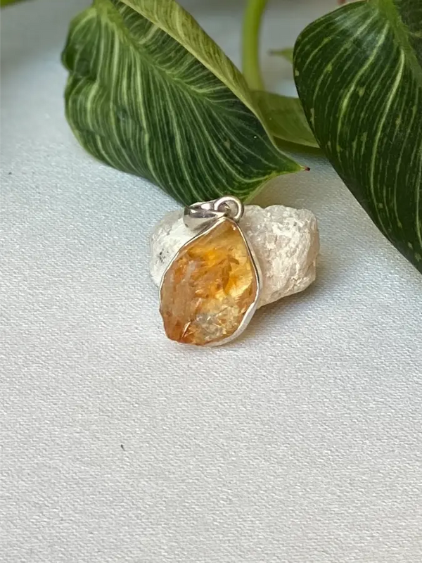 Silver pendant with a raw yellow Citrine gemstone on a textured surface with green leaves in the background