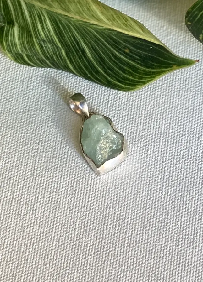 Aquamarine raw crystal stone pendant with silver setting on a textured fabric background