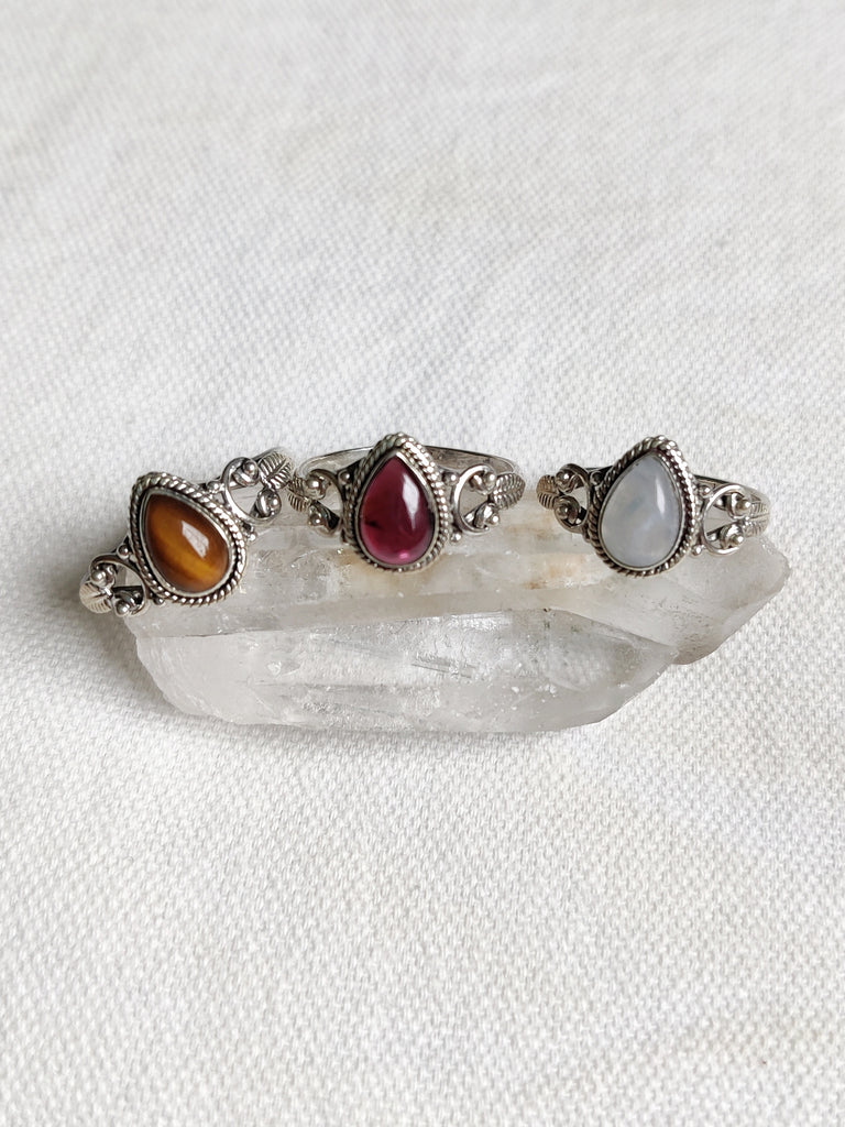 This is three sterling silver rings with a tear drop design and a crystal gemstone. One ring has a tigers eye crystal browny in colour, one has a montone crystal rainbow white in colour and one is a garnet crystal that is dark red in colour, all available at our third eye crystal and jewellery store in Auckland, New Zealand.